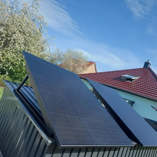 Zwei aufgeständerte Solarmodule auf einem Flachdach, dahinter ein Wohnhaus mit rotem Ziegeldach